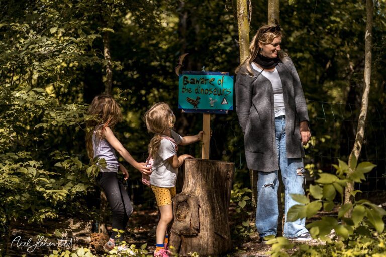 Zwei Kinder erkunden neugierig ein Schild mit der Aufschrift „Banter of the dinosaurs“ im grünen Wald – © Axel Hebenstreit