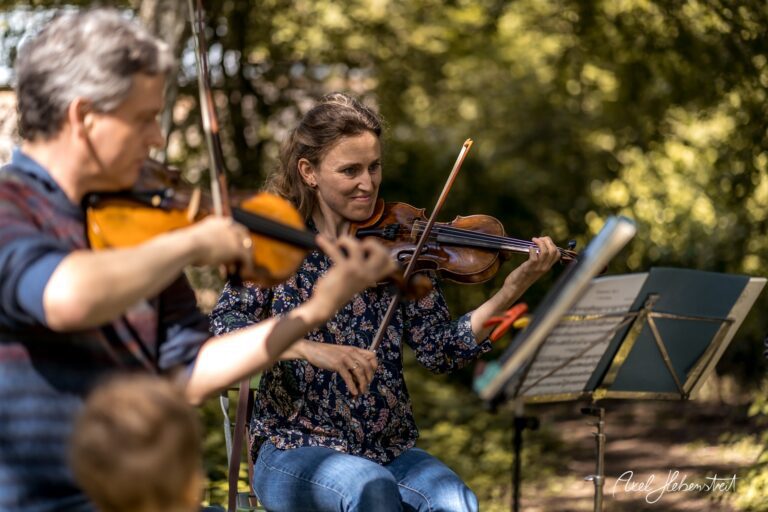 Zwei Musiker spielen im Freien Geige, umgeben von Natur und schöner Lichtstimmung – © Axel Hebenstreit.