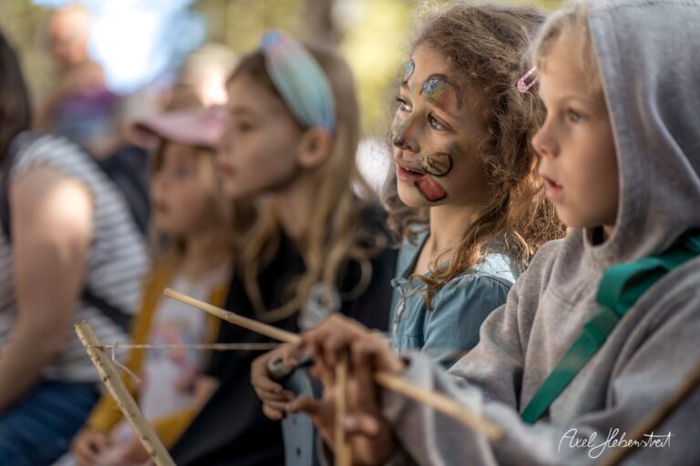 Gruppe von Kindern mit Gesichtsbemalung, die gespannt einer Aufführung im Freien zuschauen – © Axel Hebenstreit