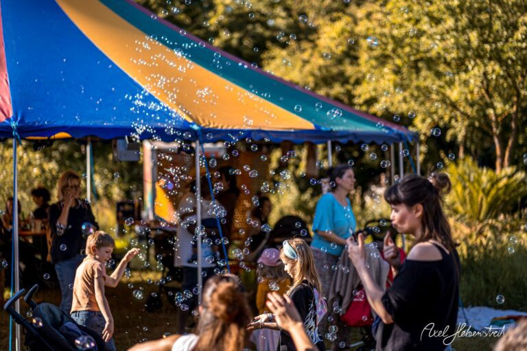 Kinder und Erwachsene spielen fröhlich mit Seifenblasen unter einem bunten Zelt auf einem Festival – © Axel Hebenstreit