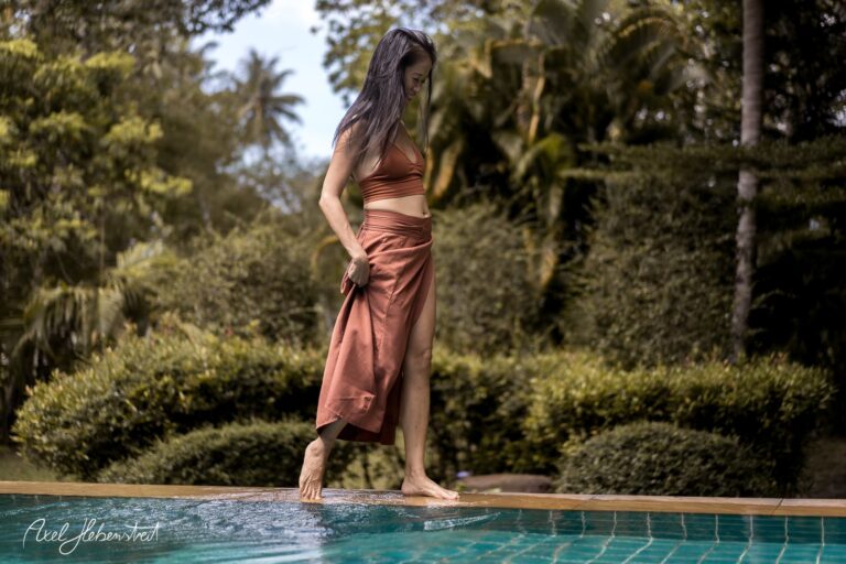 Frau in einem eleganten Outfit, die barfuß an einem Pool entlanggeht, umgeben von üppiger Natur – © Axel Hebenstreit