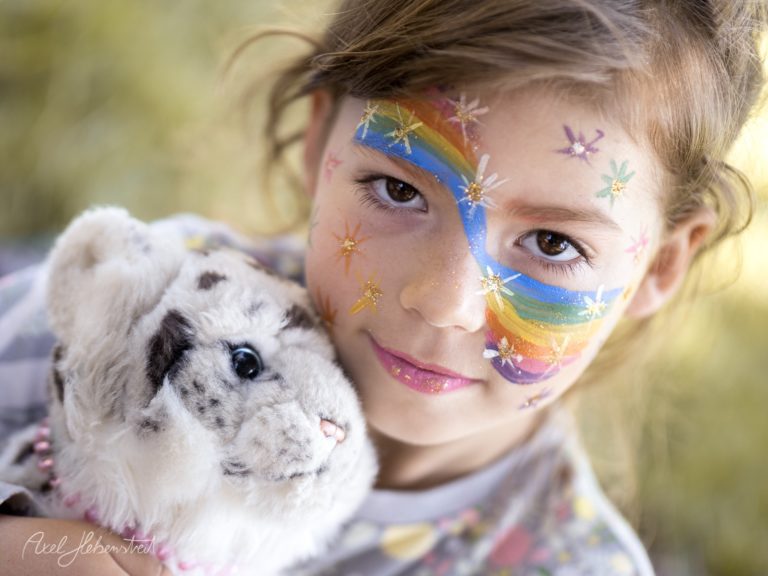 Lächelndes Kind mit buntem Regenbogen-Gesichtsbemalung hält weißes Kaninchen im Freien – © Axel Hebenstreit