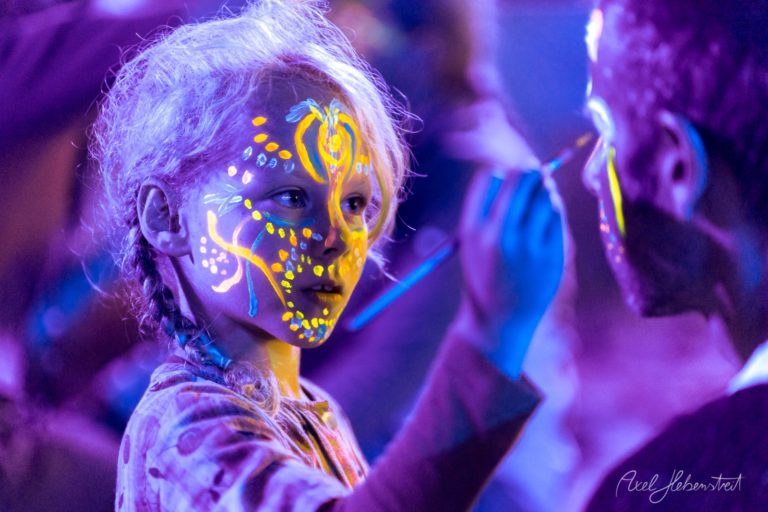 Junge Frau mit fluoreszierendem Neon-Bodypainting auf Gesicht und Körper bei Festival mit lila Atmosphäre – © Axel Hebenstreit