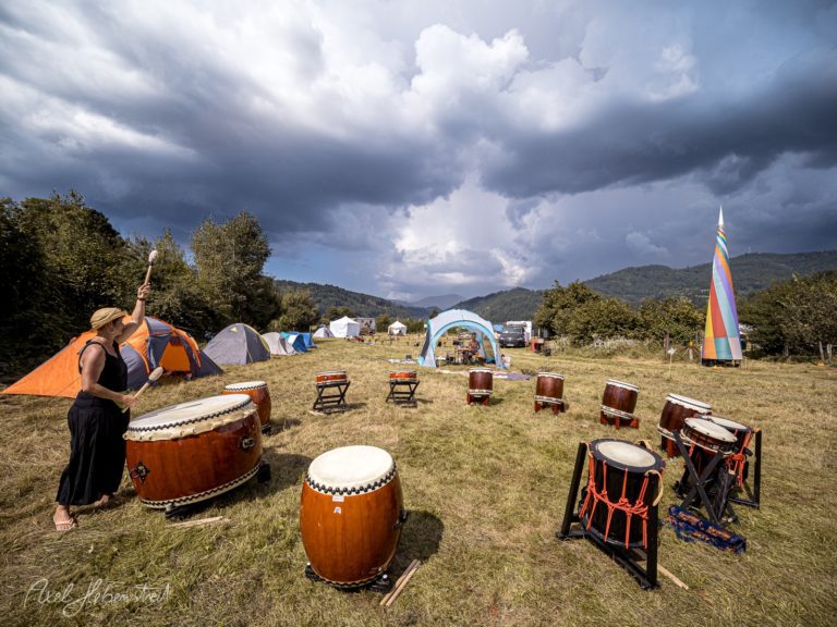 Musikerin spielt auf Handtrommel bei Outdoor-Festival mit Zelten und Bergen im Hintergrund – © Axel Hebenstreit