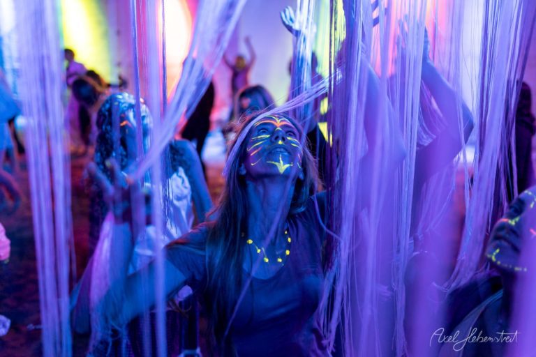 Frau mit fluoreszierendem Bodyart in lila Neonlicht bei Festival, umgeben von glühenden Lichtsäulen – © Axel Hebenstreit