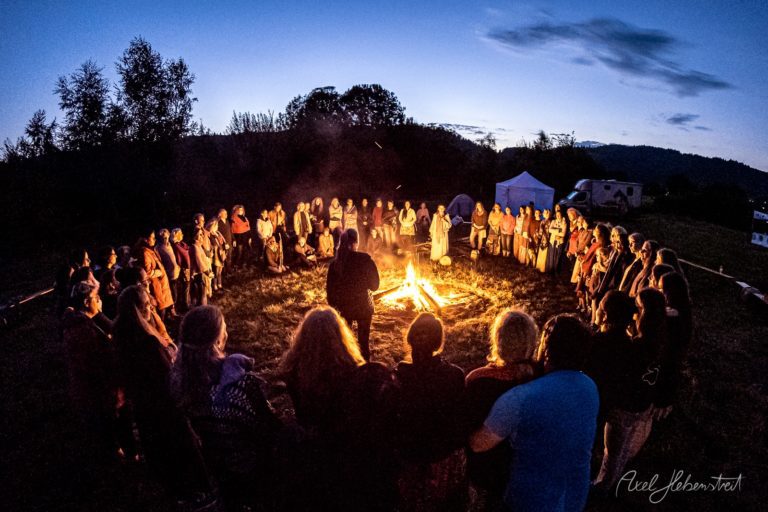 Große Gruppe von Menschen sitzt im Kreis um ein Lagerfeuer in der Dämmerung in den Bergen Bayerns – © Axel Hebenstreit