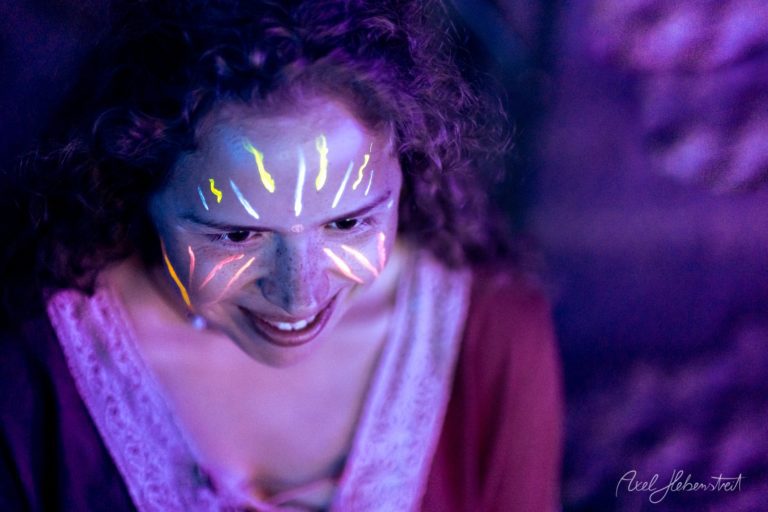 Person mit leuchtendem Neon-Bodypainting im Gesicht schaut nach unten in mystischer violetter Beleuchtung – © Axel Hebenstreit