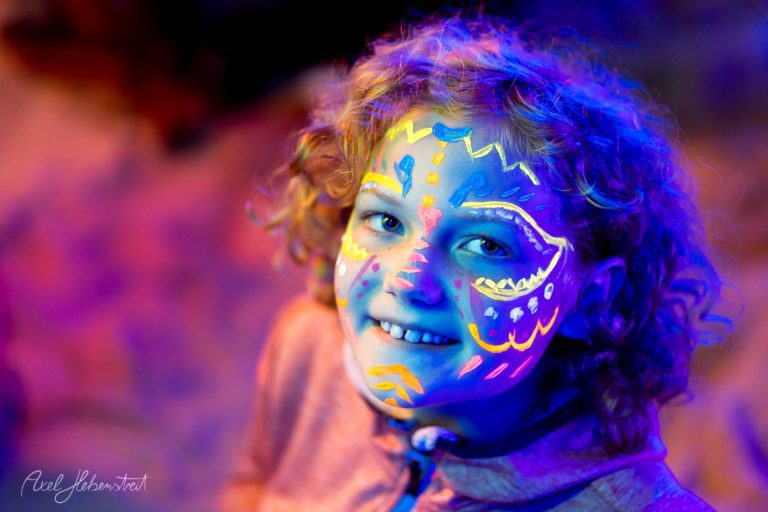Lachendes Kind mit leuchtendem Neon-Bodypainting in Blau, Gelb und Rosa vor verschwommenem buntem Hintergrund – © Axel Hebenstreit