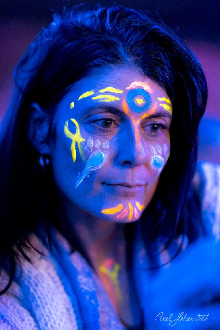 Porträt einer Frau mit fluoreszierendem Körperkunst-Design in Blau, Gelb und Orange unter UV-Licht – © Axel Hebenstreit