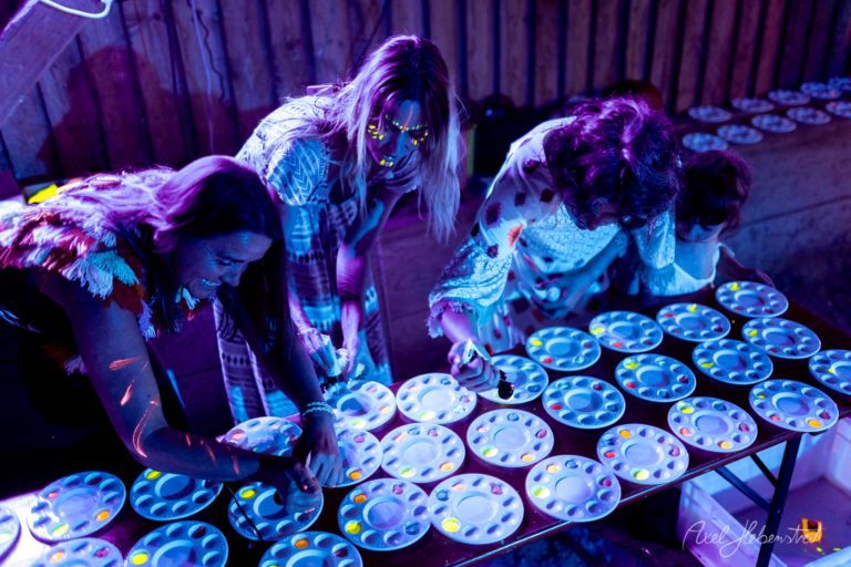 Künstler mit fluoreszierendem Body-Painting malen bei Neonlicht auf leuchtenden Paletten in einer Indoor-Installation – © Axel Hebenstreit