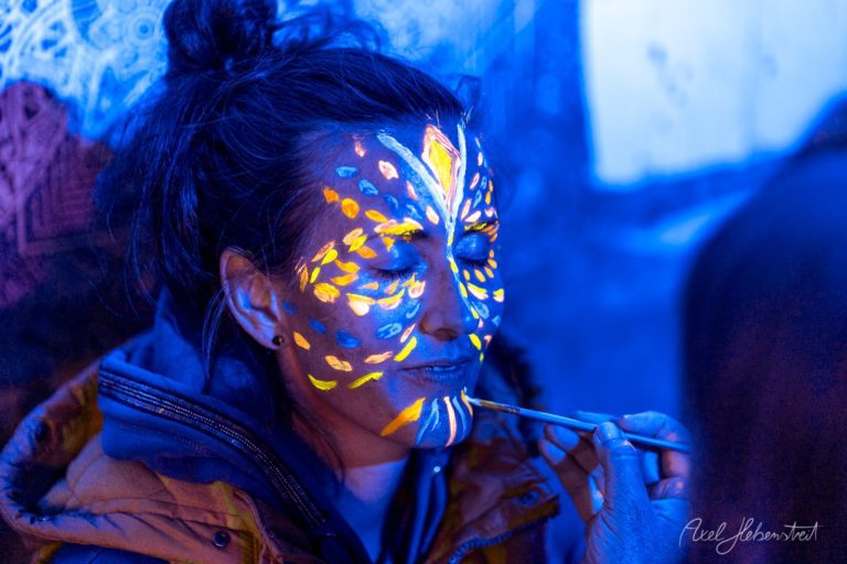 Frau mit leuchtendem Bodypainting in Blau und Gelb unter UV-Licht bei Festival – © Axel Hebenstreit