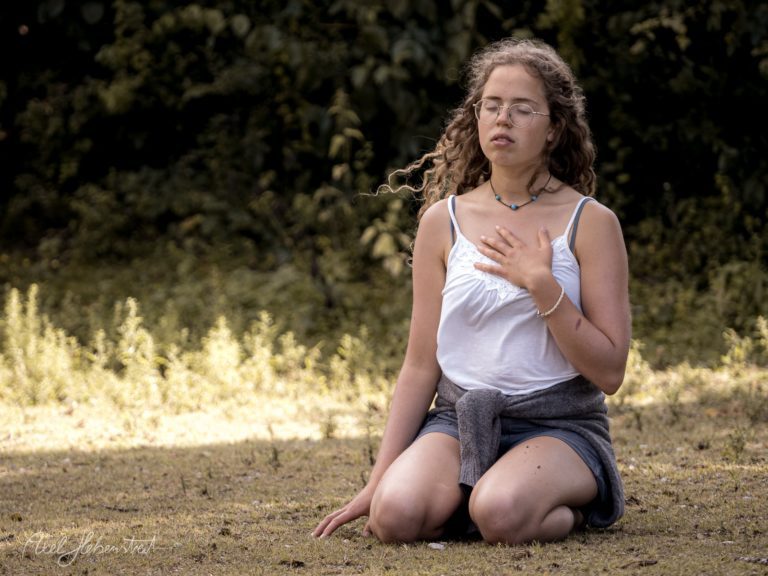 Junge Frau kniet nachdenklich in der Natur, trägt Brille und weißes Top, umgeben von grüner Vegetation – © Axel Hebenstreit