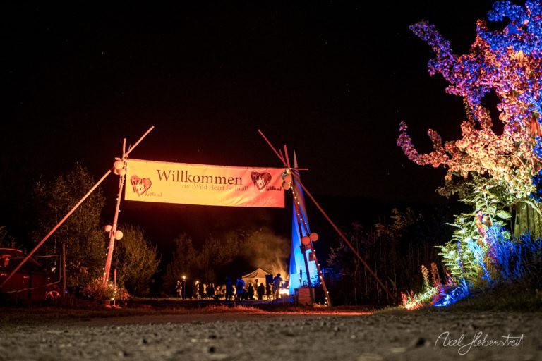Willkommen-Banner eines Festivals bei Nacht mit farbig illuminierten Blumen und Besuchern unter einem Zeltdach – © Axel Hebenstreit