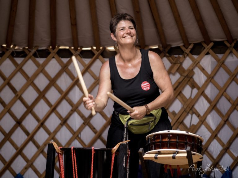Musikerin spielt lachend Schlagzeug in traditionellem Jurten-Zelt bei Live-Performance – © Axel Hebenstreit
