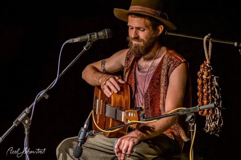 Musiker auf der Bühne sitzt mit Gitarre, trägt Hut und vermittelt eine entspannte Atmosphäre – © Axel Hebenstreit.