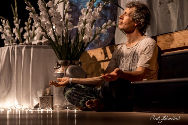 Ein meditierender Mann sitzt in einer ruhigen, stimmungsvollen Umgebung zwischen Kunstblumen und sanften Lichtern – © Axel Hebenstreit