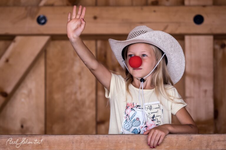 Ein fröhliches Mädchen mit einer roten Clownsnase und einem Hut winkt von einer Holzplattform aus – © Axel Hebenstreit