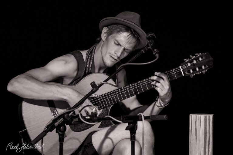 Ein Musiker sitzt konzentriert mit einer Gitarre auf der Bühne und spielt leidenschaftlich – © Axel Hebenstreit