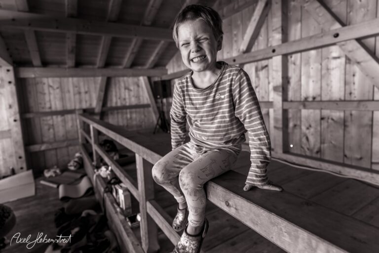 Ein lächelndes Kind sitzt auf einer Holzplattform in einer rustikalen Umgebung. – © Axel Hebenstreit