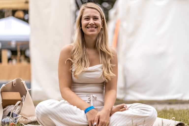 Lächelnde junge Frau im entspannten Sitz auf einem Festivalgelände, umgeben von Zeltständen. – © Axel Hebenstreit