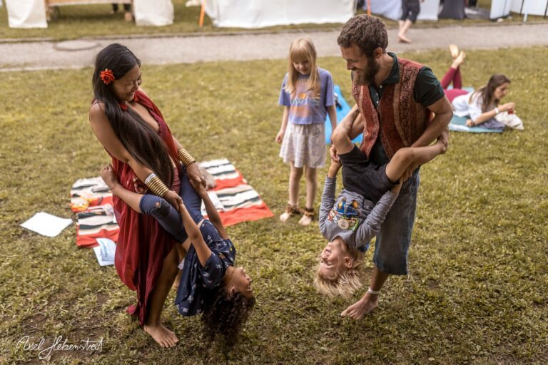 Zwei Erwachsene heben lachende Kinder in der Luft, während im Hintergrund andere Menschen auf einem Festival entspannen – © Axel Hebenstreit