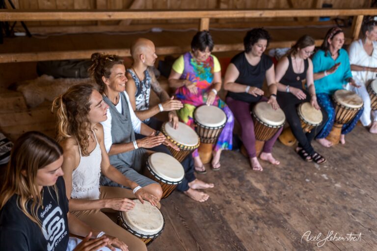 Eine Gruppe von Menschen spielt gemeinsam Trommeln in einer warmen und einladenden Holzhalle – © Axel Hebenstreit