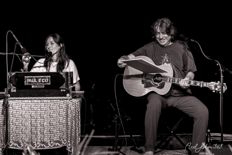 Ein Musiker mit Gitarre und eine Sängerin am Harmonium strahlen während eines Konzerts Freude und Harmonie aus – © Axel Hebenstreit
