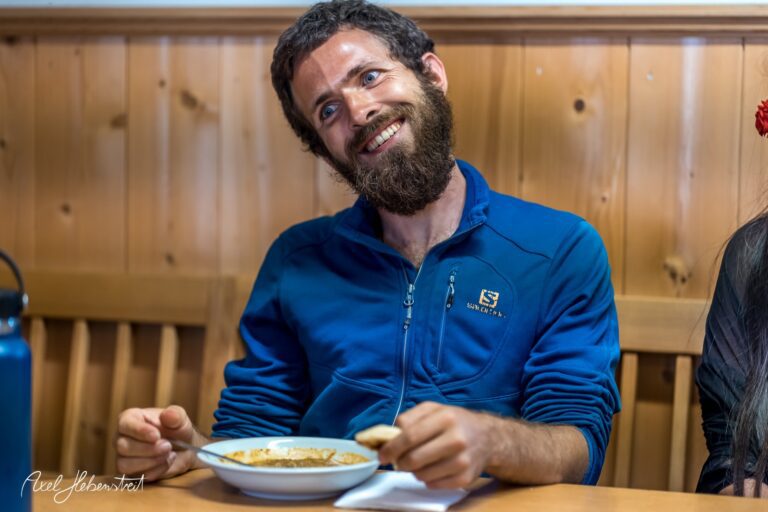 Lächelnder Mann mit Bart in blauer Jacke sitzt an einem Tisch und genießt sein Essen – © Axel Hebenstreit