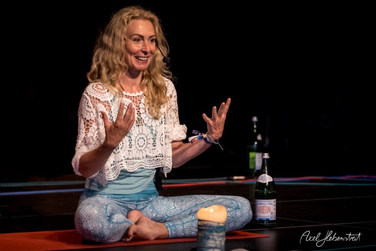 Eine Frau in Yogakleidung sitzt lächelnd im Licht, während sie motivierend spricht, umgeben von Yoga-Equipment – © Axel Hebenstreit.