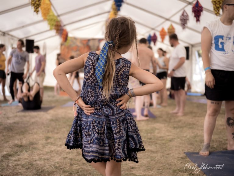 Ein Mädchen in einem bunten Kleid steht entspannt in einem Yogazelt voller Menschen – © Axel Hebenstreit.