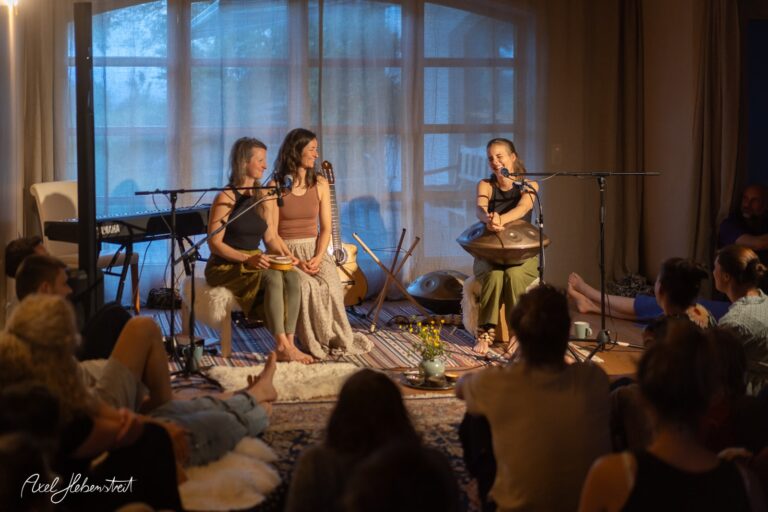 Drei Musikerinnen mit Handpan und Gitarre bei einem intimen Wohnzimmerkonzert vor Publikum in warmer Abendstimmung.