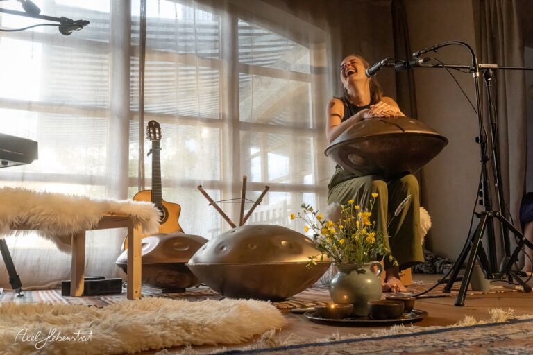 Musikerin spielt Handpan und singt in sonnendurchflutetem Raum mit Gitarre und Blumen bei Yoga-Session
