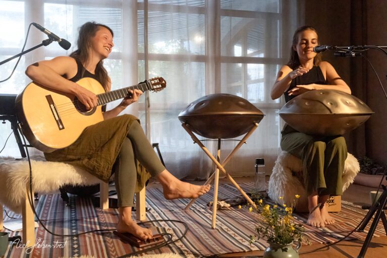 Zwei Musikerinnen musizieren entspannt mit Gitarre und Handpan in hellem, lichtdurchflutetem Raum.