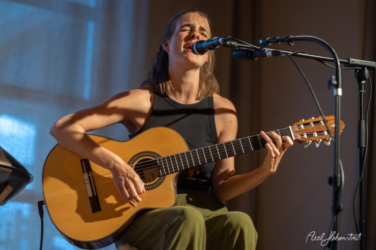 Musikerin singt emotional mit Akustikgitarre vor Mikrofon in stimmungsvollem Bühnenlicht
