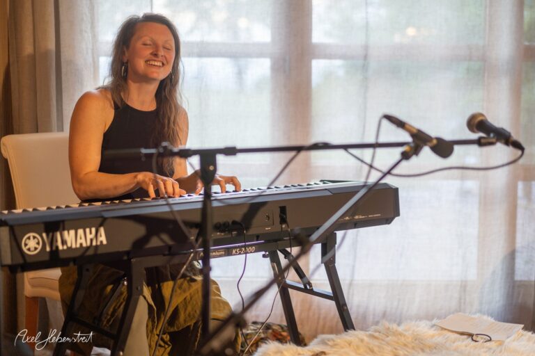 Musikerin spielt lächelnd am Keyboard in hellem Raum mit großen Fenstern