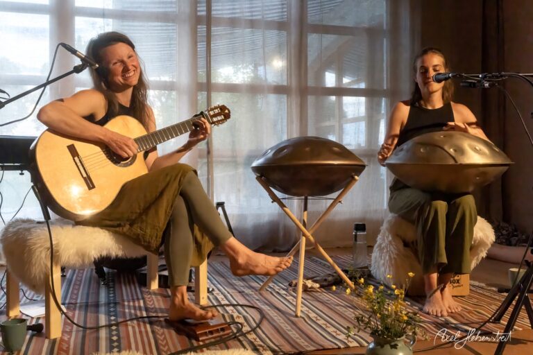 Zwei Musikerinnen spielen Gitarre und Handpan in hellem Raum mit großen Fenstern bei Live-Performance.