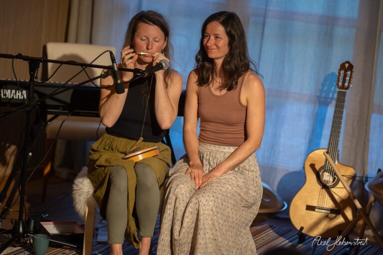 Zwei Musikerinnen bei intimem Konzert, eine spielt Mundharmonika am Mikrofon, Yamaha-Keyboard und Gitarre im Hintergrund.