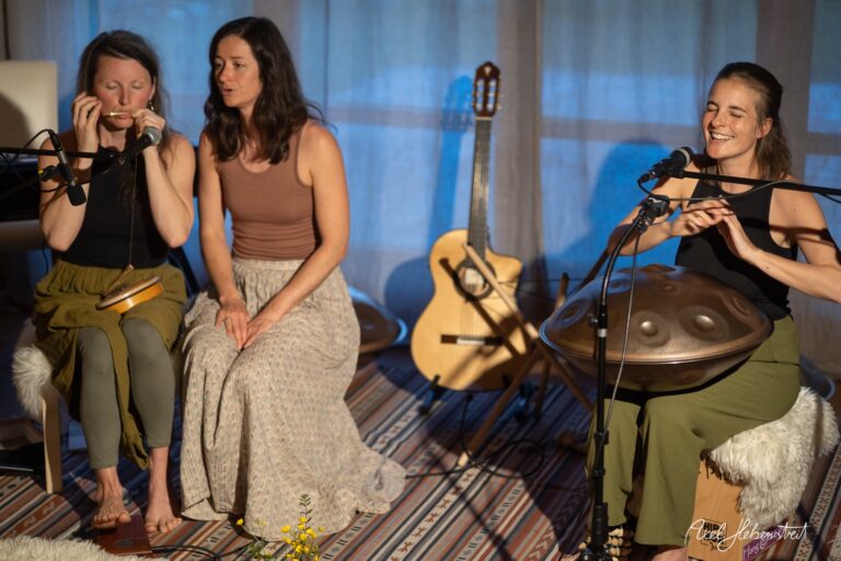 Drei Musikerinnen bei einer intimen Aufnahme-Session mit Hang-Drum, Mundharmonika und Gitarre in gemütlichem Ambiente.