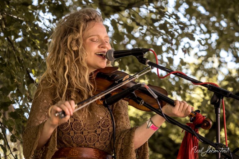 Eine Musikerin mit Geige singt leidenschaftlich unter einem Baum bei einem Festival – © Axel Hebenstreit