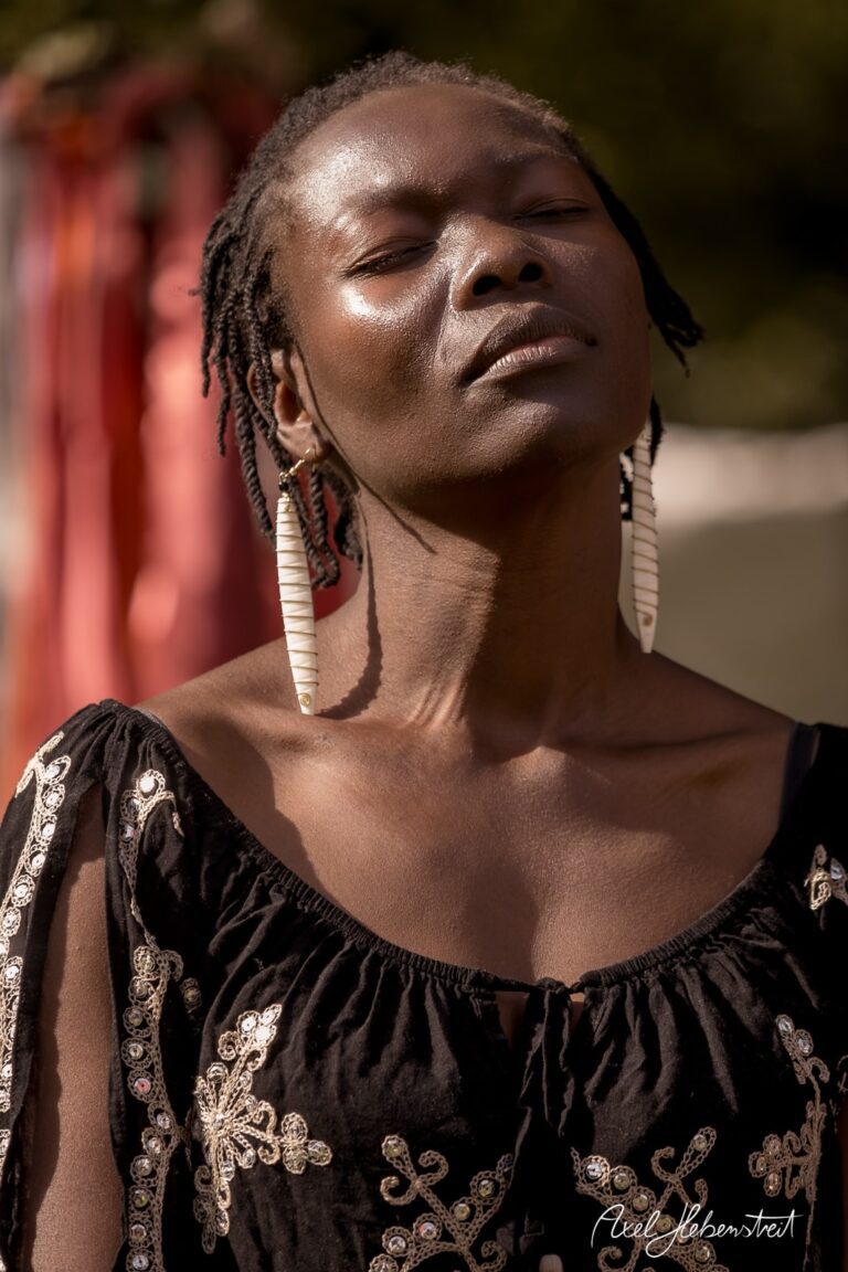 Eine Frau mit Dreadlocks und großen Ohrringen genießt das Sonnenlicht in einem festlichen, farbenfrohen Ambiente – © Axel Hebenstreit