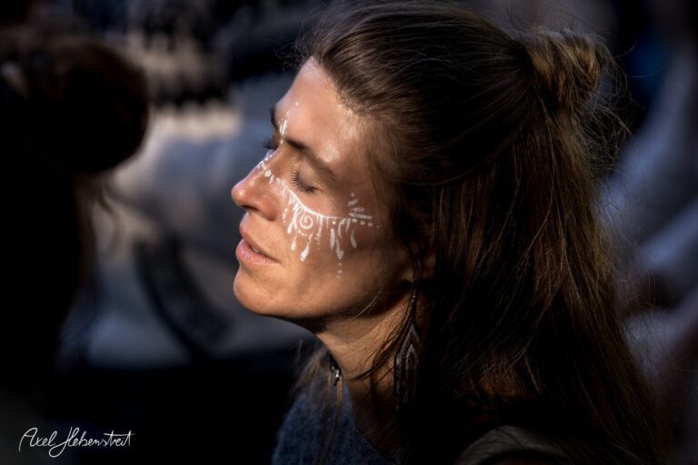 Eine Frau mit auffälligem Gesichtsbemalung meditiert in einer entspannten Festivalatmosphäre – © Axel Hebenstreit