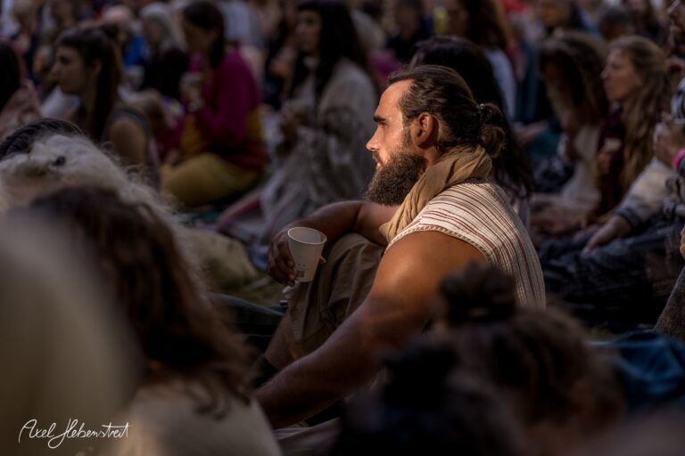 Ein nachdenklicher Mann mit Bart sitzt in einer Gruppe von Festivalbesuchern und hält einen Becher in der Hand – © Axel Hebenstreit