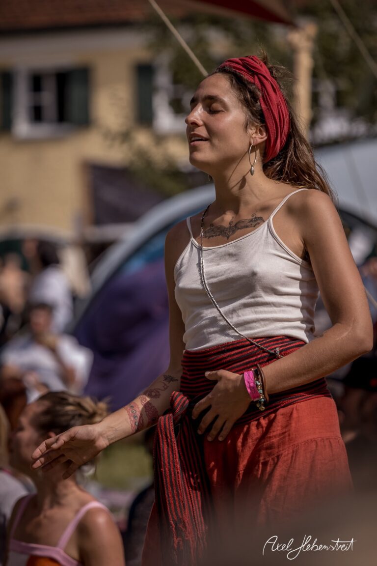 Frau in entspannter Pose mit roten Tuch und Tattoos, umgeben von festivalartigen Kulissen – © Axel Hebenstreit