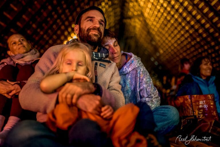 Eine Familie sitzt entspannt und zufrieden bei einem Konzert, umgeben von warmem Licht und harmonischer Stimmung – © Axel Hebenstreit