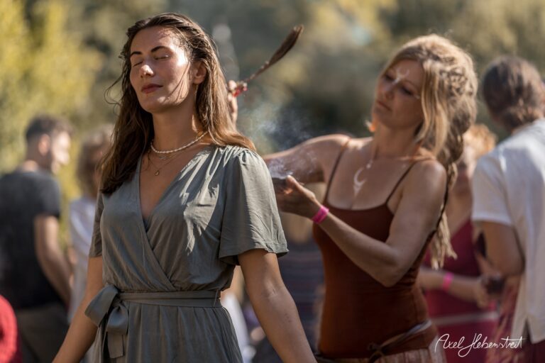 Zwei Frauen in entspannter Atmosphäre bei einem Festival, eine wird mit Rauchzeremonie verwöhnt – © Axel Hebenstreit