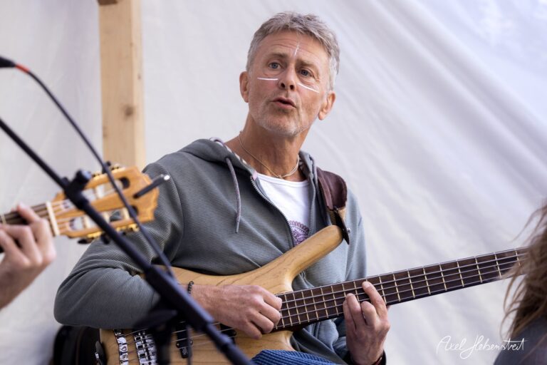 Musiker spielt Bassgitarre auf einer kleinen Bühne, umgeben von einer entspannten Festivalatmosphäre – © Axel Hebenstreit
