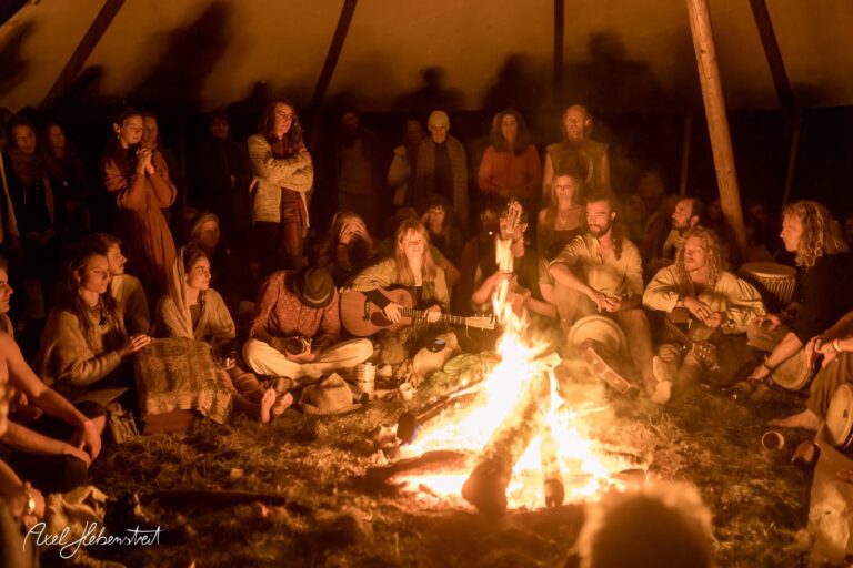 Eine Gruppe von Menschen sitzt um ein Lagerfeuer, singt, musiziert und genießt die gemeinschaftliche Atmosphäre. – © Axel Hebenstreit