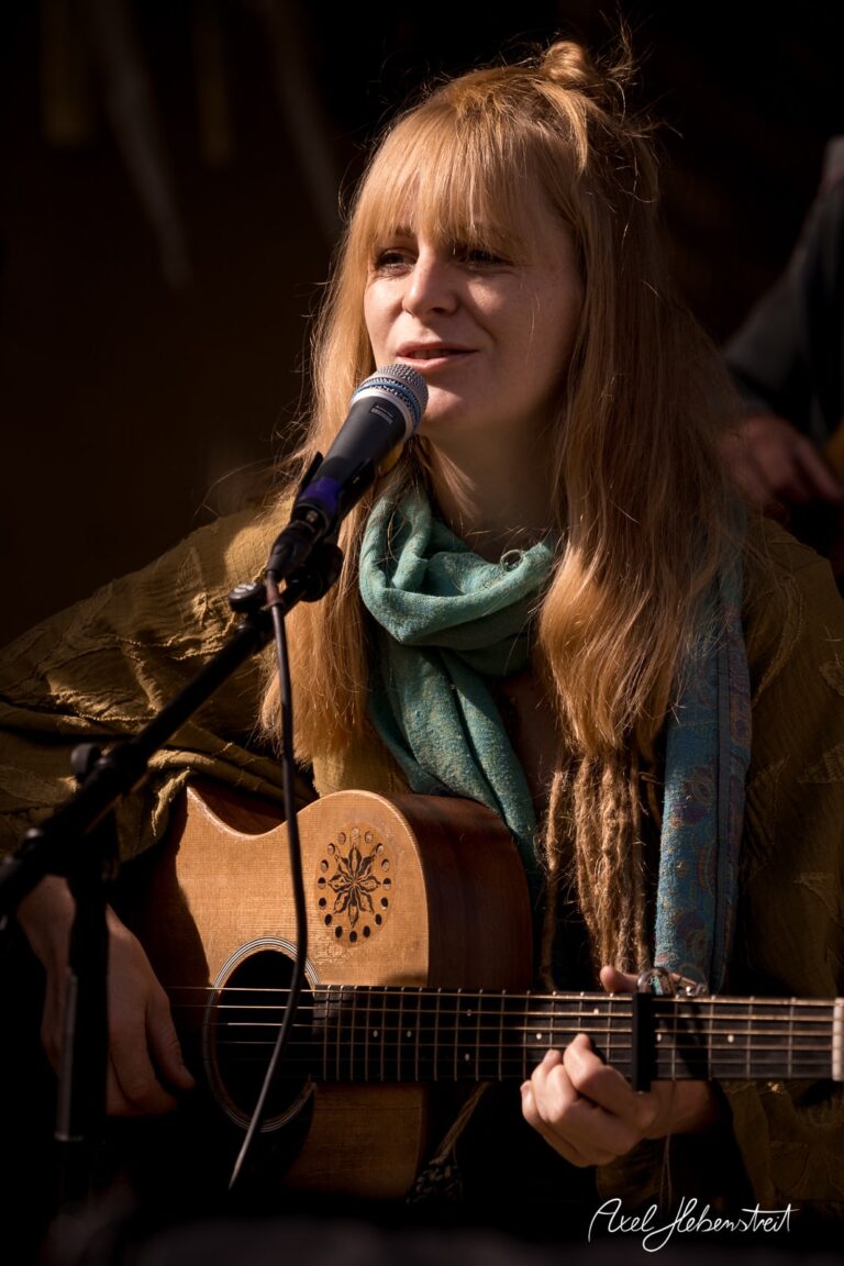 Frau mit Gitarre singt begeistert und strahlt Freude aus, umgeben von einer entspannten Festivalatmosphäre. – © Axel Hebenstreit