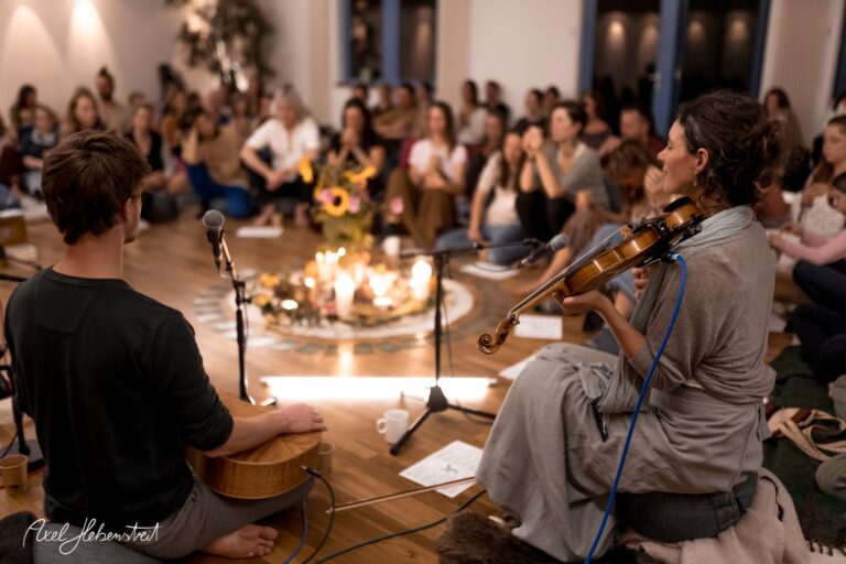 Eine Gruppe von Menschen lauscht einer energetischen Musiksitzung im gemütlichen Ambiente eines großen Raums – © Axel Hebenstreit.