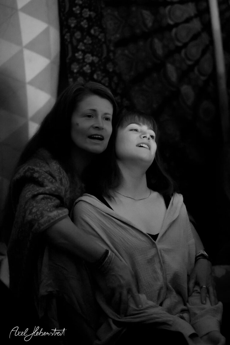 Zwei Frauen, die emotional miteinander verbunden sind, umgeben von einem farbenfrohen und stimmungsvollen Hintergrund. – © Axel Hebenstreit